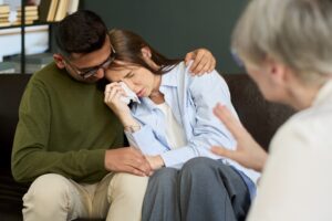 A distraught woman wipes away tears while being comforted by a partner during a counseling session. A counselor sits nearby, speaking with the couple, illustrating the emotional impact of a traumatic event or loss.